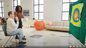 A woman helping a man seated in a chair throw a ball. 
